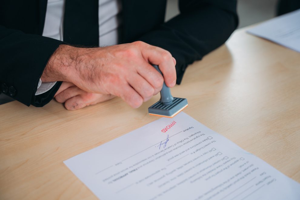 Businessman hand stamping an approved stamp on a text document at a table with a contract form paper in the background.